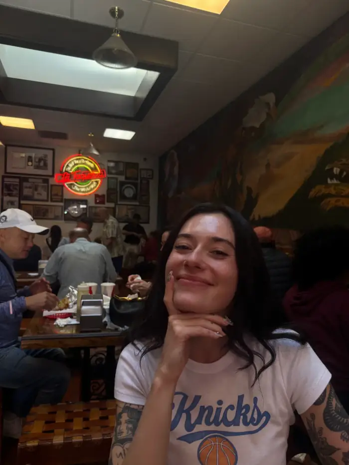 olivia in a knicks shirt in a busy diner, smiling with her chin on her hand