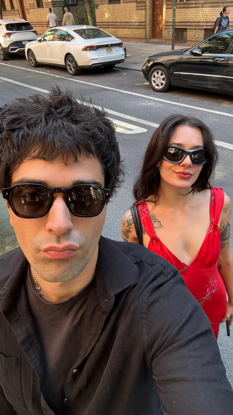 thomas in sunglasses on the sidewalk, olivia a step behind him, traffic past them